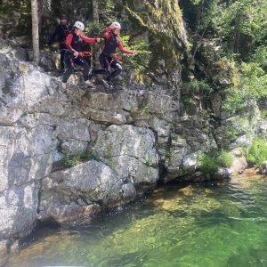 Trois personnes équipées de casques et baudriers sautant depuis une falaise rocheuse vers une eau é饰.